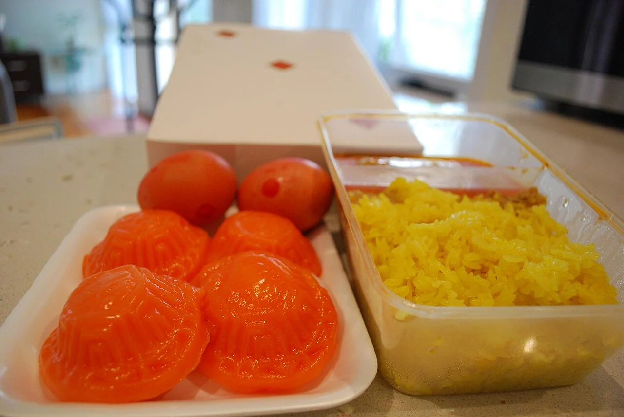 A white plate holds four shiny orange ang ku kueh, with two red eggs and a box of yellow rice nearby.