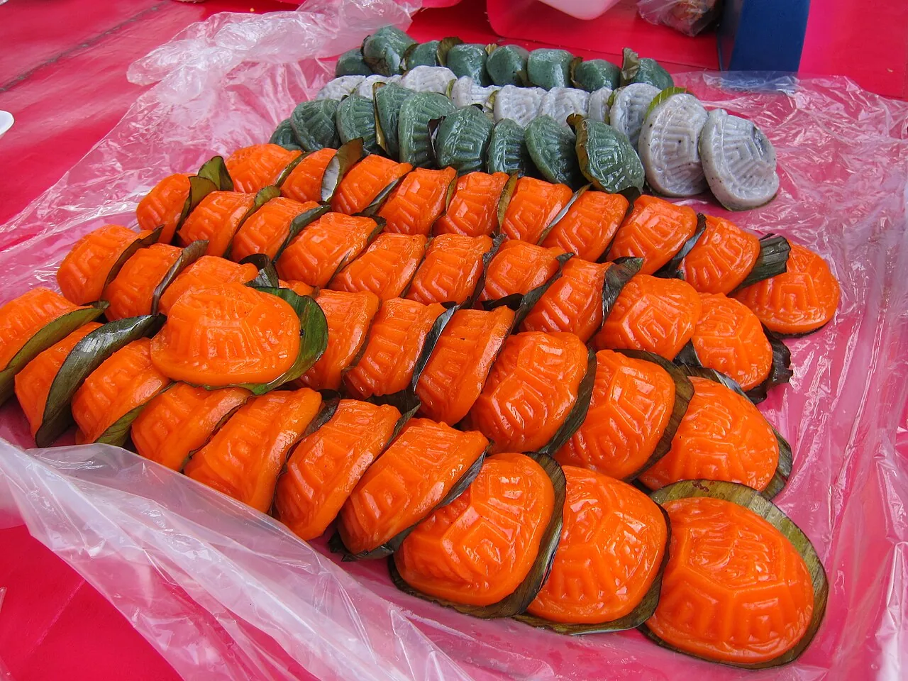 Many bright orange ang ku kueh with leaf bases are arranged on plastic, with some green and white ones behind.