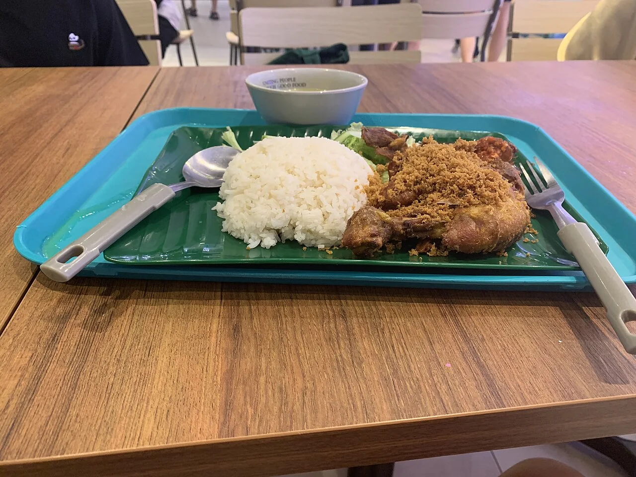 A plate on a green tray holds white rice, fried chicken topped with crumbs, and a spoon and fork beside a small bowl.