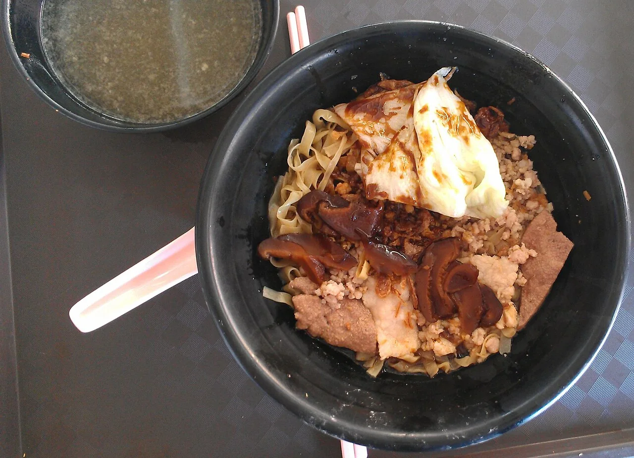 A black bowl of noodles topped with a fried egg, sliced mushrooms, and minced meat sits beside a bowl of soup.