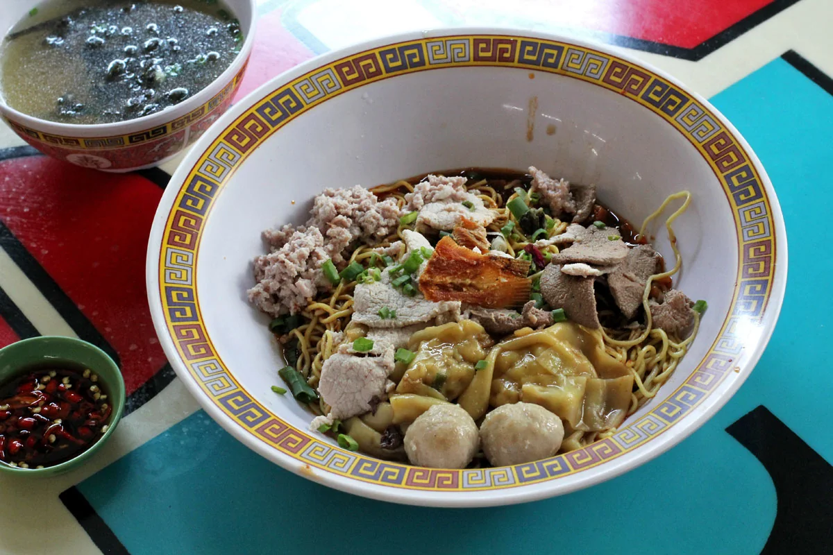 A patterned bowl of noodles with minced meat, sliced liver, meatballs, and dumplings sits beside a bowl of soup and a small dish of chopped chilli.
