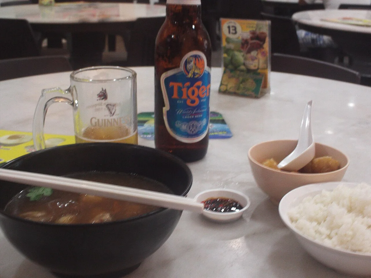 A black bowl of bak kut teh soup with chopsticks sits beside rice and a small sauce dish.
