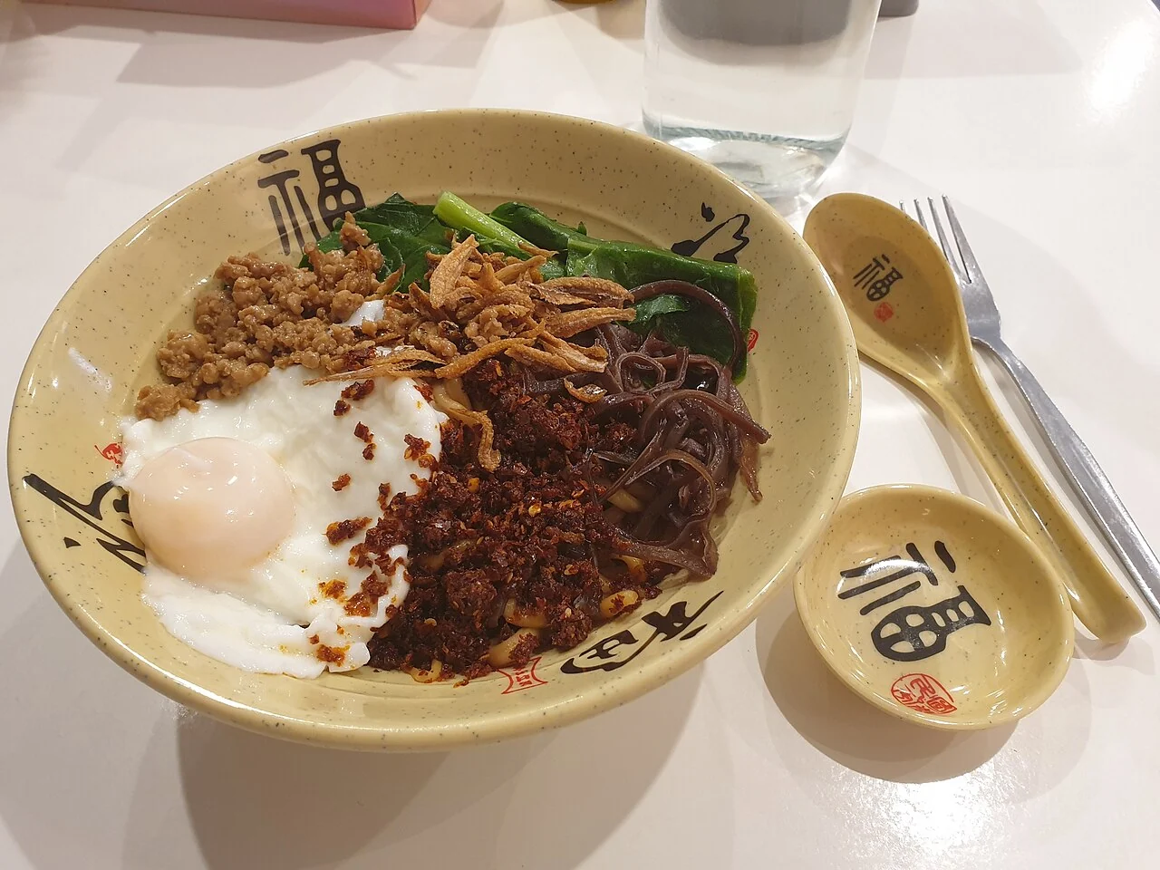 A bowl of noodles has an egg, green vegetables and brown toppings, with a spoon resting at the side.