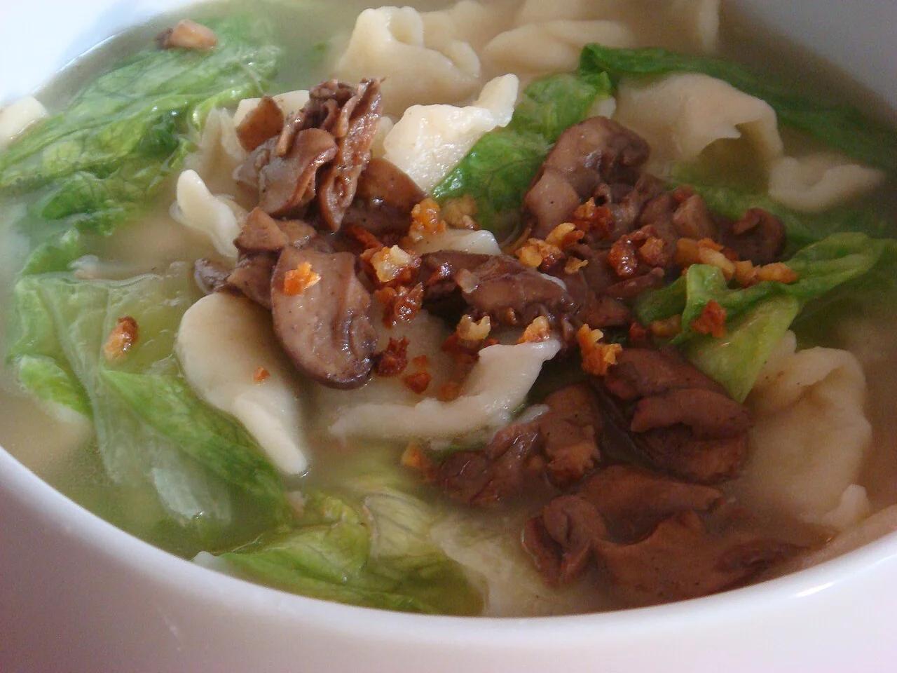 Wide pieces of noodles sit in soup with green leaves, mushrooms and crisp brown toppings.