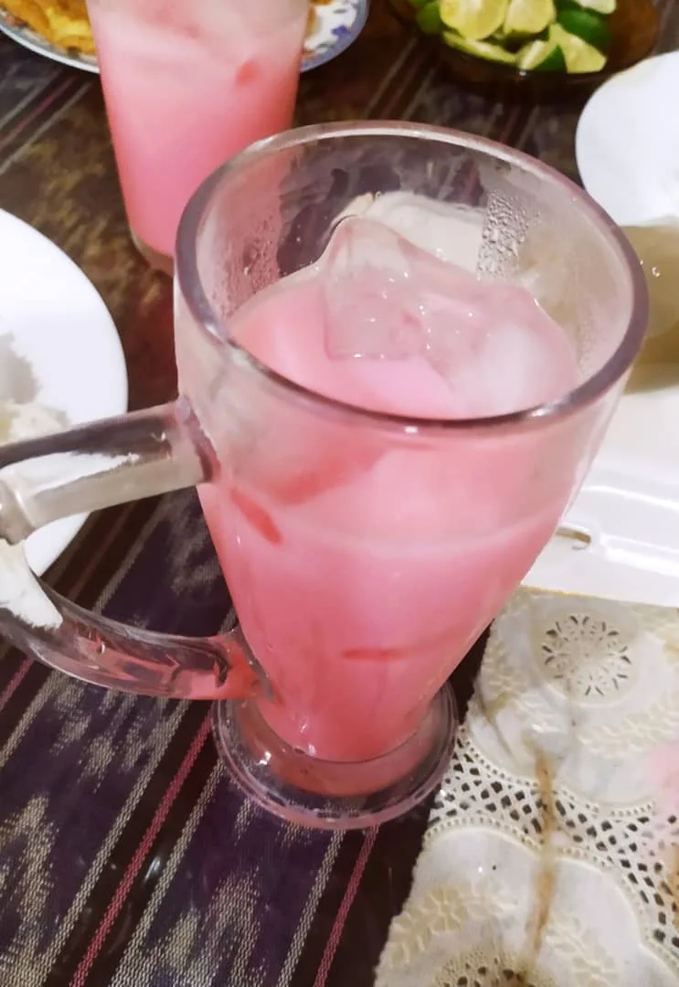 A glass mug filled with pink iced drink and ice cubes rests on a striped tablecloth.