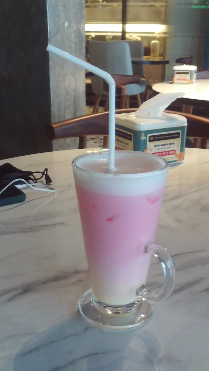 A tall glass of pink iced milk drink with a white straw sits on a marble table beside a tissue box.