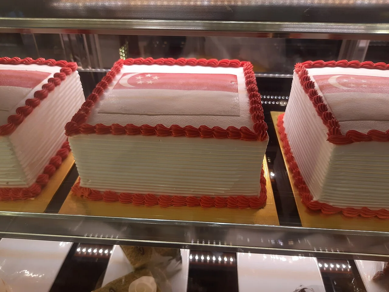 Three rectangular white cakes decorated with red piping and a Singapore flag design sit on a glass shelf.