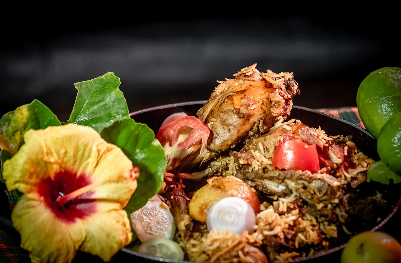 A dark bowl holds spiced rice with a large piece of chicken, tomato, onion, lime, and a yellow flower.