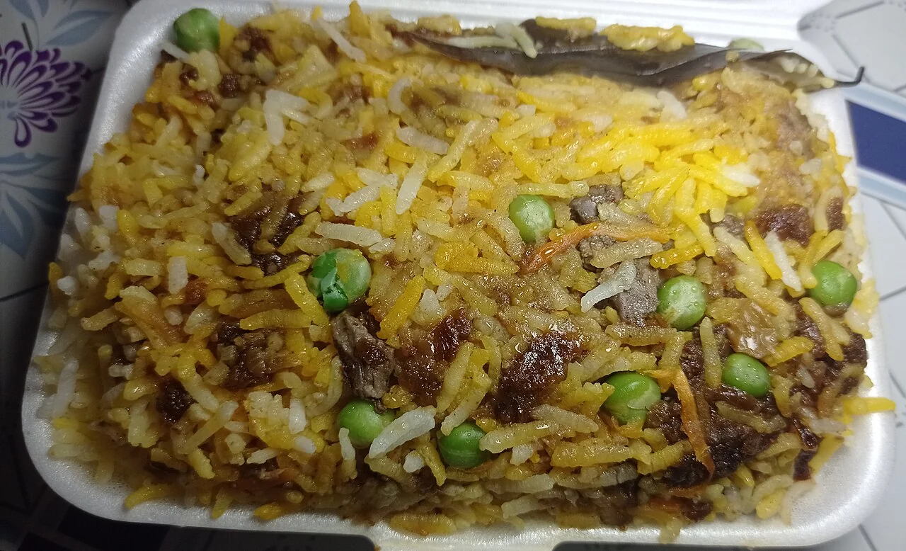 Yellow and white rice with green peas, dark meat pieces, and a leaf on top sits in a white takeaway box.