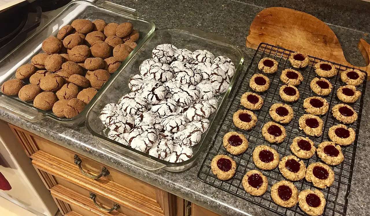 Brown cookies, sugar-dusted cookies and cookies with red jam centres are arranged on dishes and a wire rack.