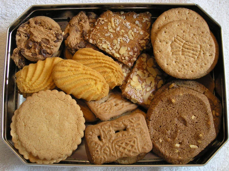 An open metal tin contains an assortment of cookies, including round, rectangular, and ridged varieties.