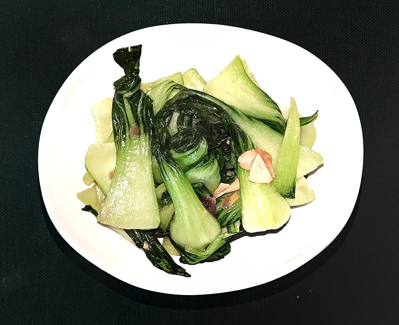 A white plate holds cooked bok choy with pale green stems and dark green leaves, along with sliced mushrooms and ginger.