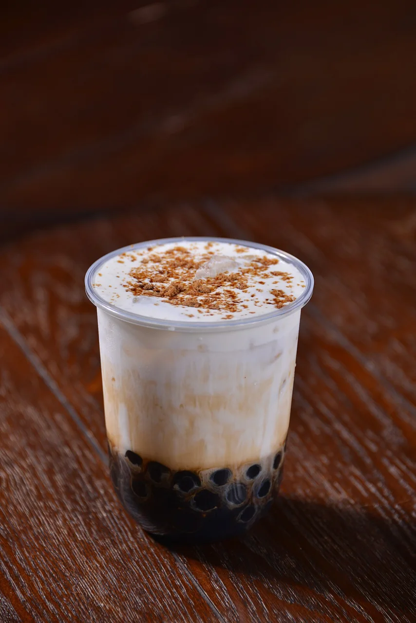 A clear plastic cup of milk tea with dark tapioca pearls at the bottom is topped with foam and brown powder, resting on a wooden table.