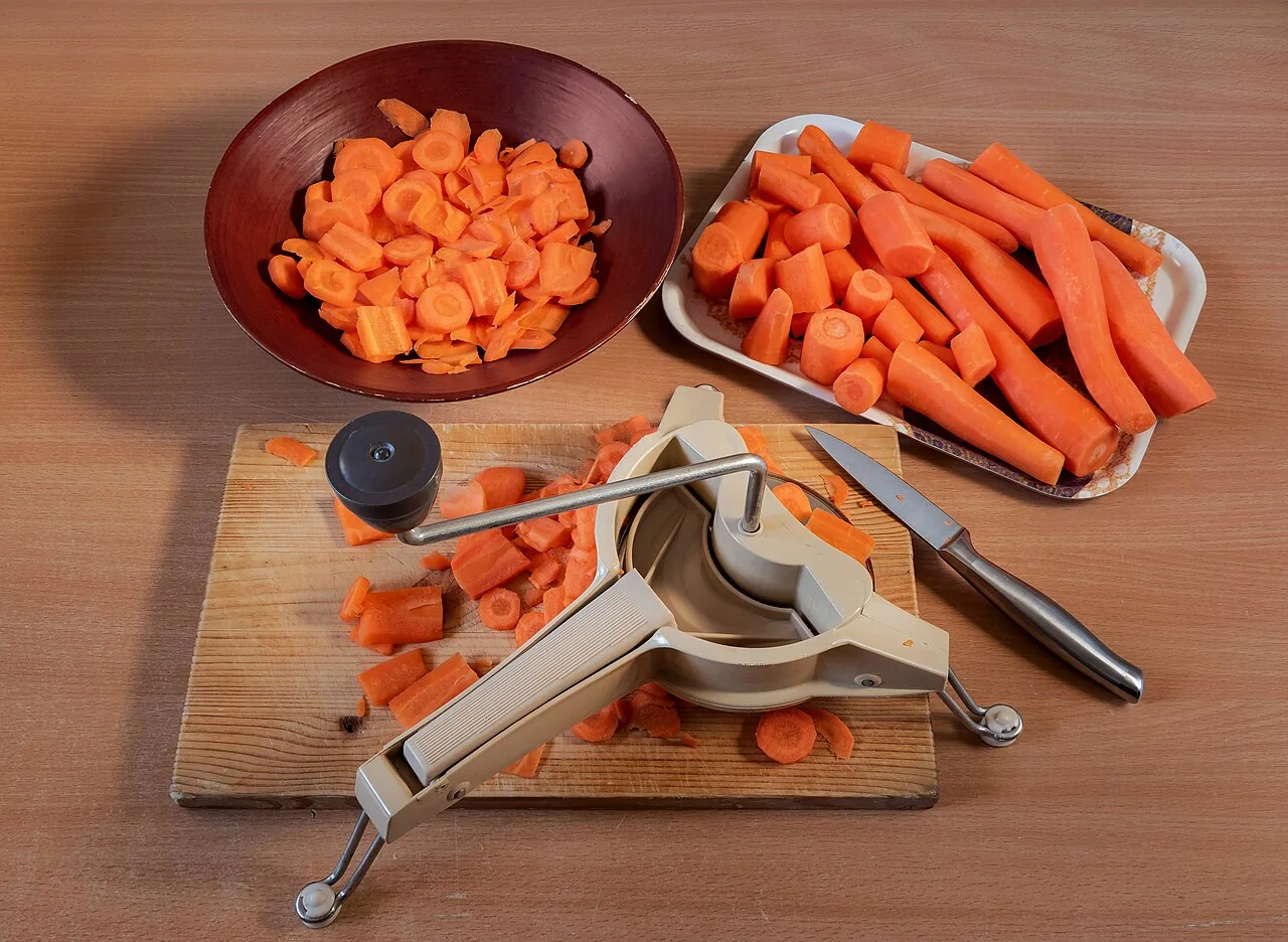 Sliced and cut carrots are arranged in a bowl and on a tray next to a slicer on a wooden board.