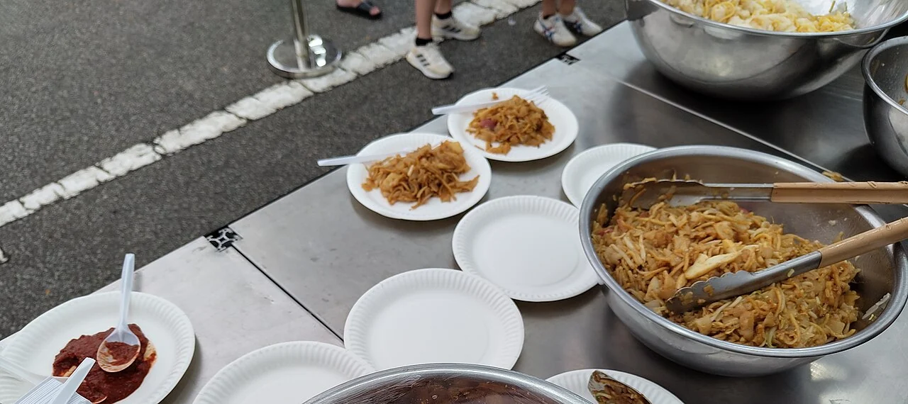 Small portions of fried noodles sit on white paper plates beside a large metal bowl of noodles and a spoon of chilli.