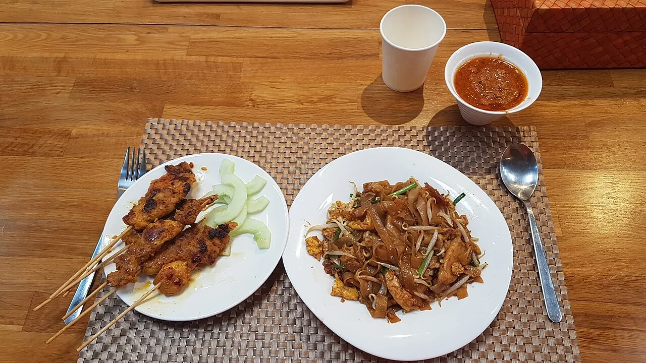 Flat rice noodles with bean sprouts sit on a white plate beside skewers of grilled meat and cucumber slices.