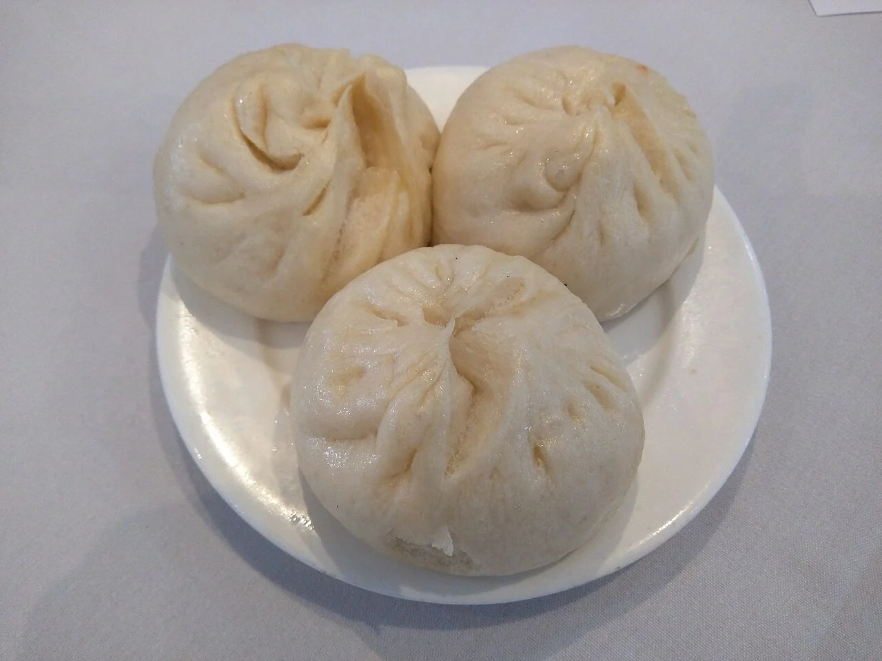 Three white steamed buns with pleated tops sit together on a small white plate.