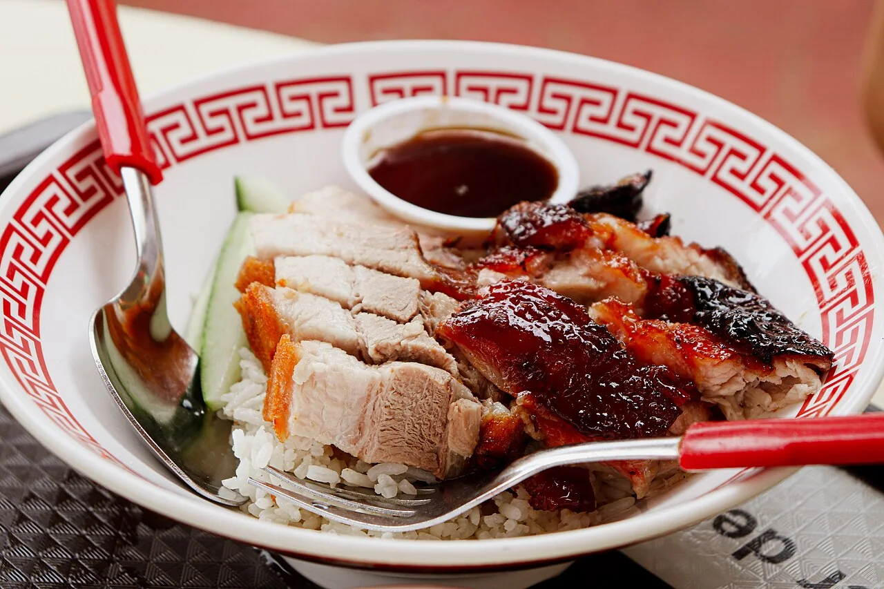A bowl of rice topped with sliced char siew and roasted pork, served with a small dish of sauce.