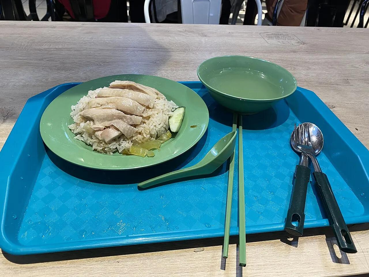 Sliced chicken rests on a mound of rice on a green plate, with a bowl, spoon, and chopsticks on a blue tray.