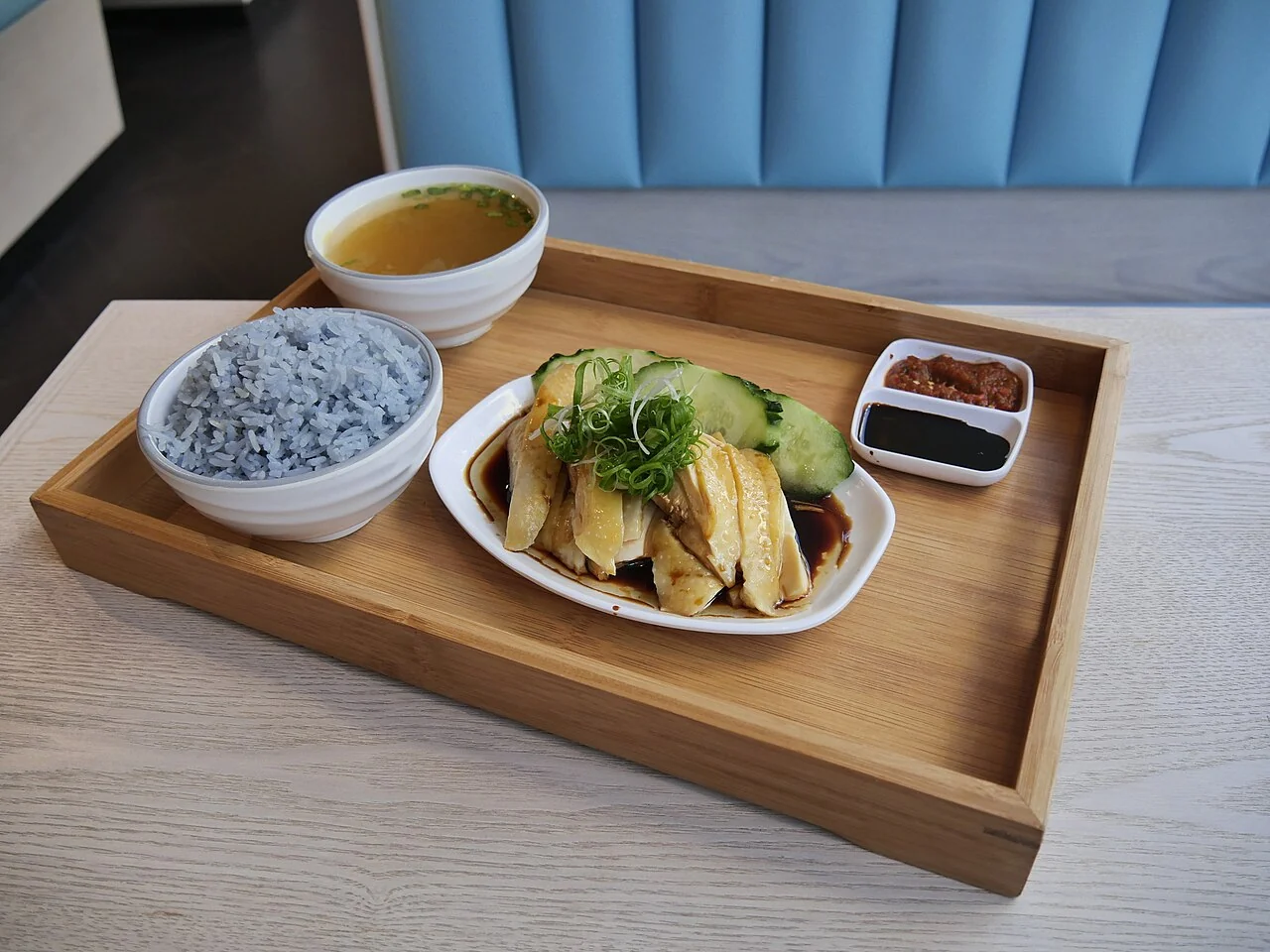 Sliced chicken with soy sauce and scallions sits on a white plate next to blue rice and a bowl of soup.