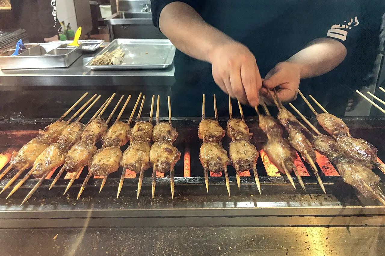 A person uses both hands to turn chicken wings on skewers over a long, glowing grill.