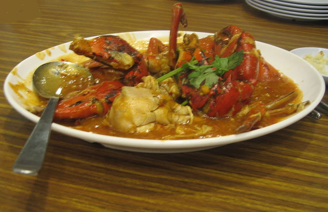 A white plate holds red crab pieces in thick orange chilli sauce, topped with a sprig of coriander.