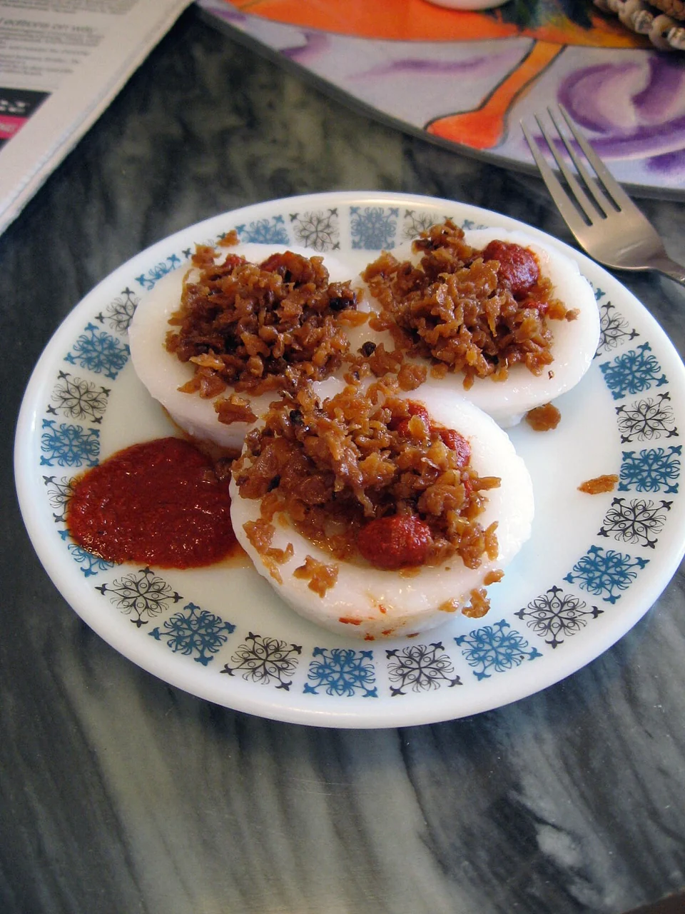 Three soft white steamed rice cakes on a patterned plate, topped with brown preserved radish and red chilli sauce.