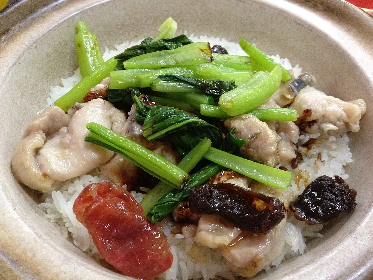 Rice in a clay pot is topped with pale chicken pieces, green vegetables and a red sausage slice.