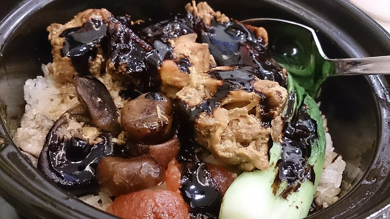 A dark clay pot holds rice topped with glossy dark meat, mushrooms, sausage slices and green leafy vegetables.