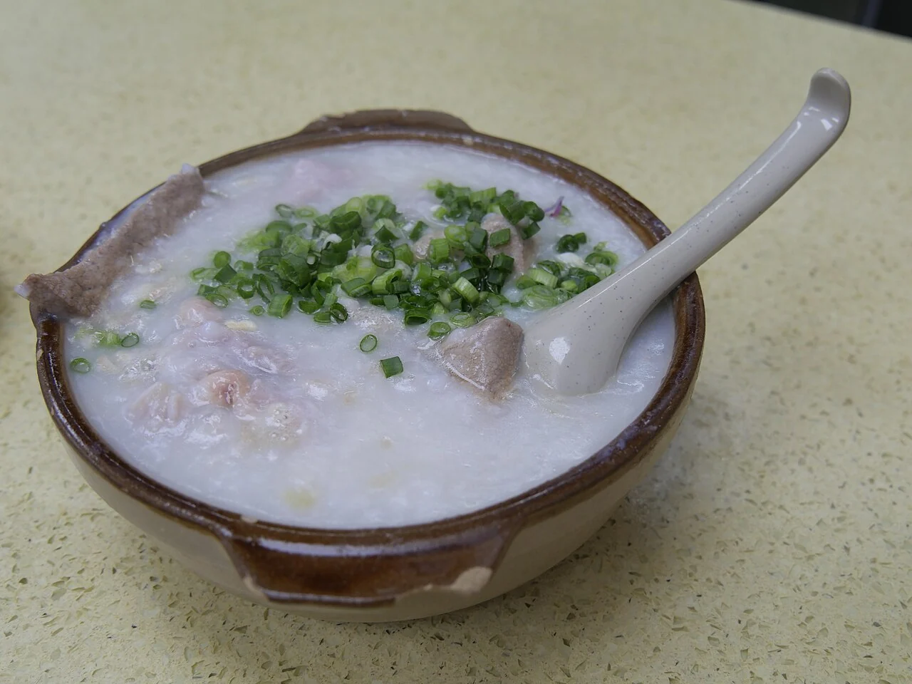 A brown bowl of white rice porridge is topped with chopped green spring onions and pieces of meat, with a spoon inside.