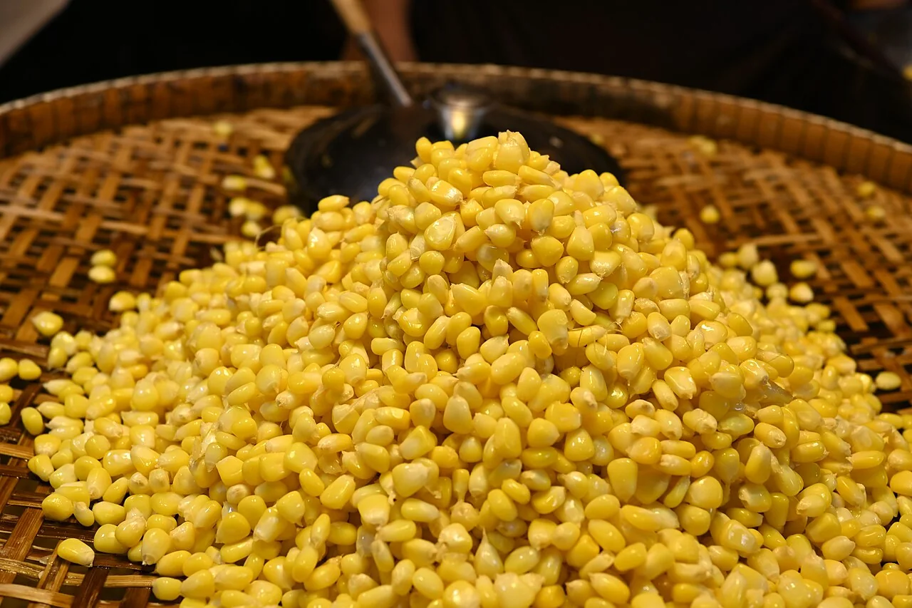 A heaping pile of fresh yellow corn kernels sits on a woven bamboo tray with a metal spoon.
