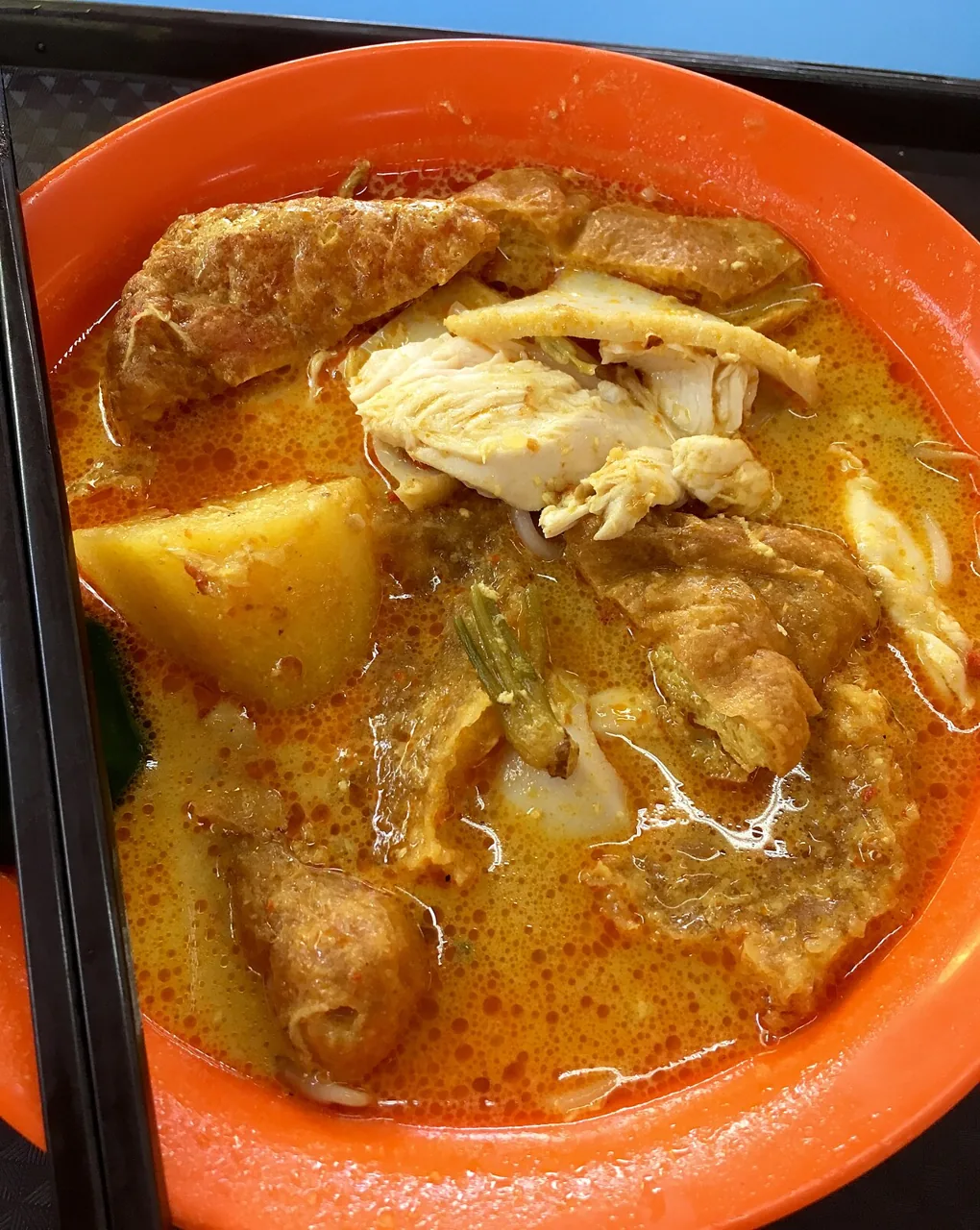 A bowl of curry noodle soup holds pieces of chicken, potato and fried tofu puffs in orange coconut gravy.