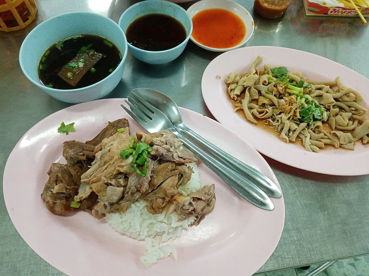 A pink plate of white rice topped with sliced braised duck and spring onions, with soup and noodles nearby.