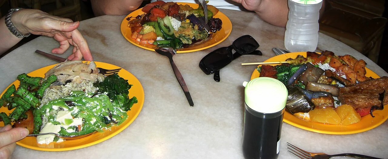 Plates of rice with green vegetables and colourful side dishes share a table with drinks.