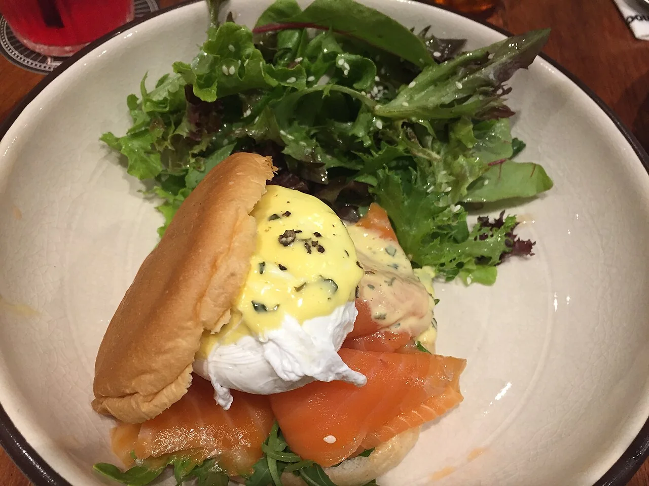 A poached egg with sauce sits on smoked salmon and a bun beside a green salad.