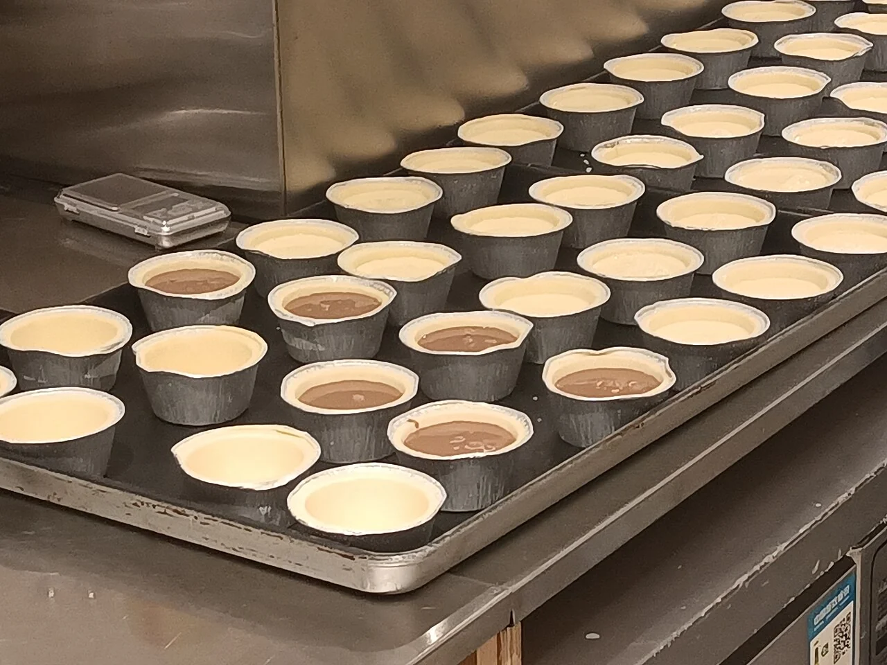 Rows of small tart shells on a metal tray are filled with pale yellow and brown custard mixtures.