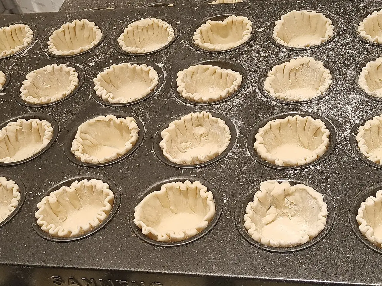 Many pale, fluted pastry shells sit in the cups of a dark baking tray dusted with flour.