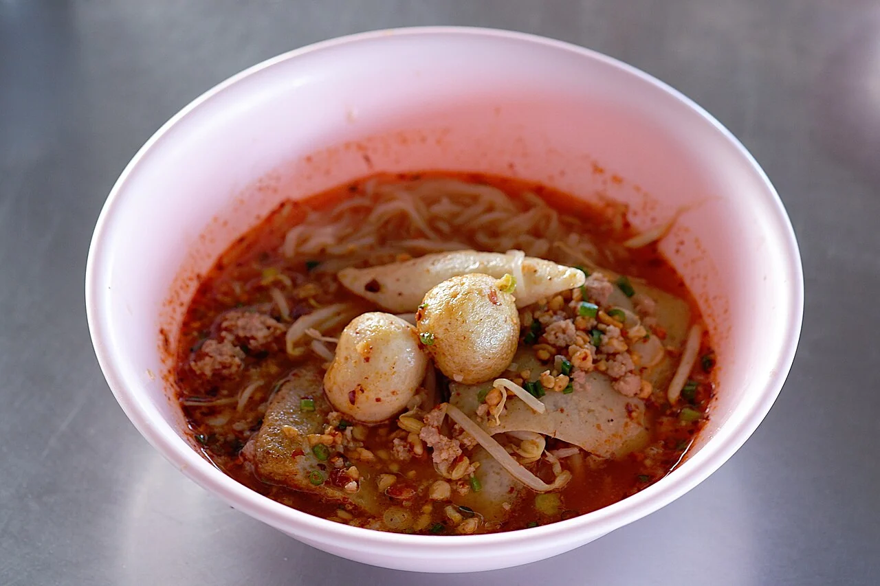 A pink bowl of noodles in red tom yum soup with fish balls, minced meat, and bean sprouts.
