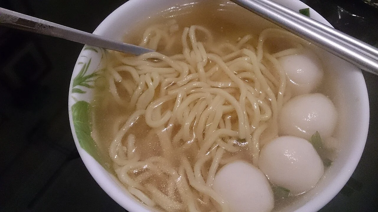 White fish balls and yellow noodles sit in clear broth in a white bowl, with chopsticks resting on the rim.