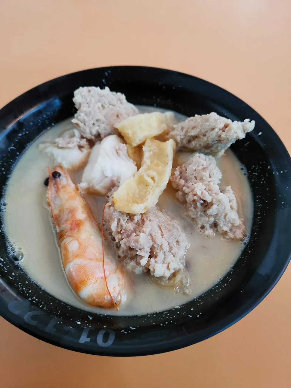 A large prawn rests across a black bowl of pale soup, with pieces of fish and other toppings.