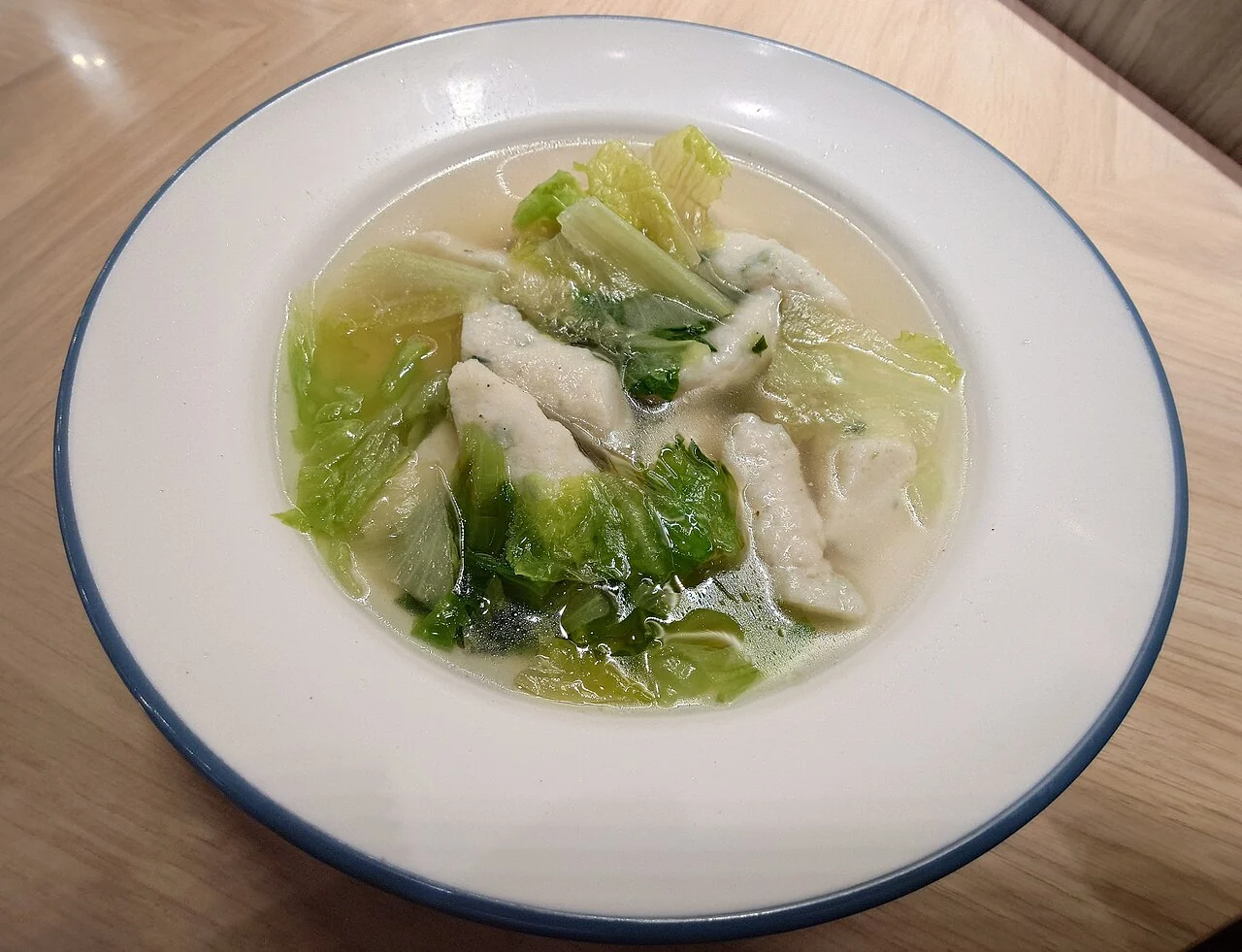 A white bowl with a blue rim holds clear soup, pale fish pieces and green lettuce leaves on a wooden table.