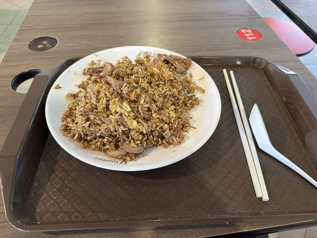 A white plate of fried rice with egg and onions sits on a brown tray with chopsticks and a spoon.