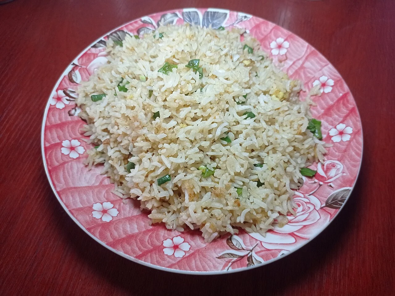 A pink floral plate holds a mound of fried rice mixed with chopped green onions on a red table.