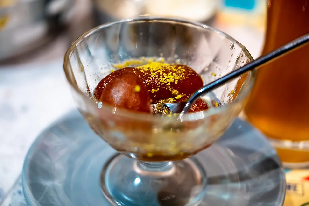 Two gulab jamun in a glass bowl topped with yellow crumbles and a spoon.