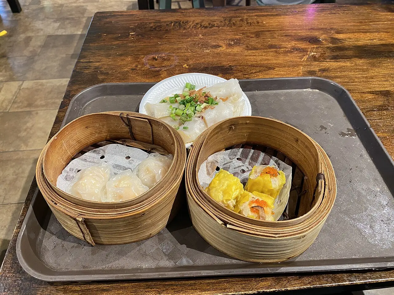 Two bamboo steamers hold dumplings on a metal tray, with a plate of rice noodle rolls topped with green onions.