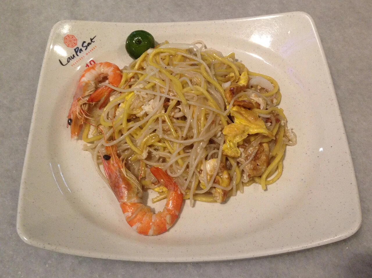 A plate of yellow noodles with two large prawns, bean sprouts, egg and a green lime half.