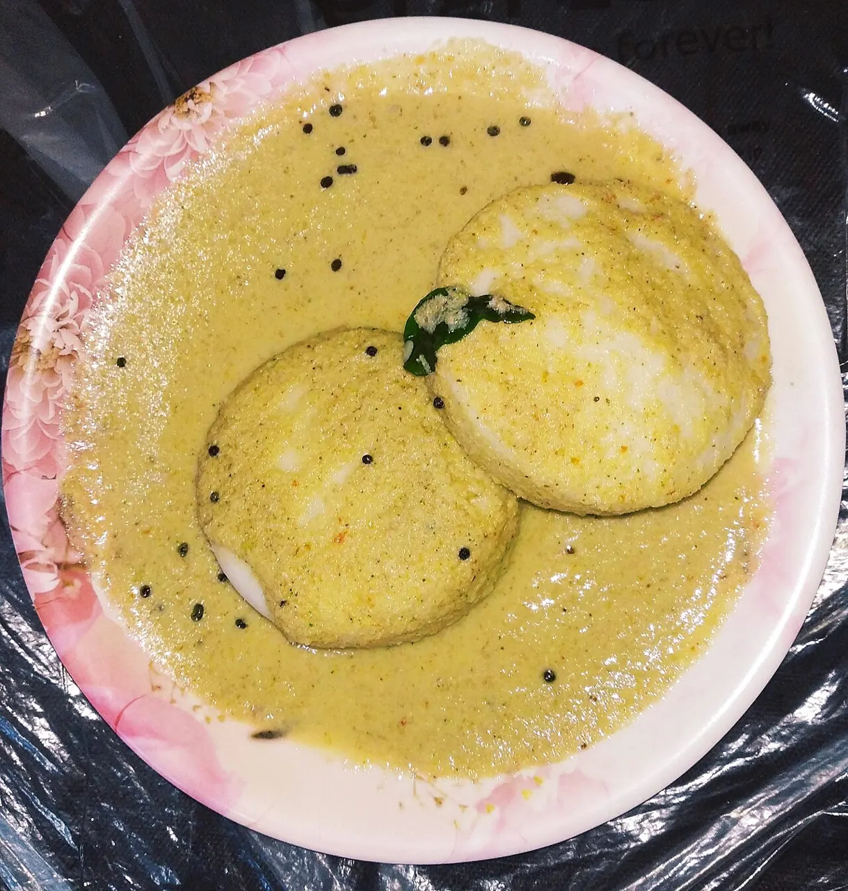 Two round white idli sit on a pink floral plate, covered in a thick green chutney with mustard seeds.