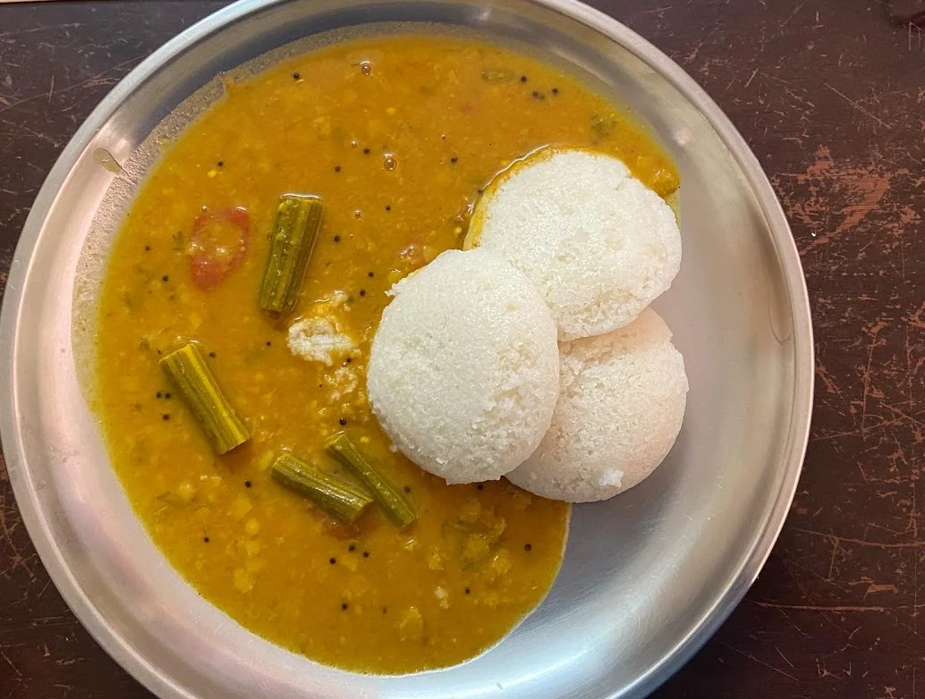 Three white idli rest in a bowl of yellow sambar, which contains visible pieces of green drumstick.