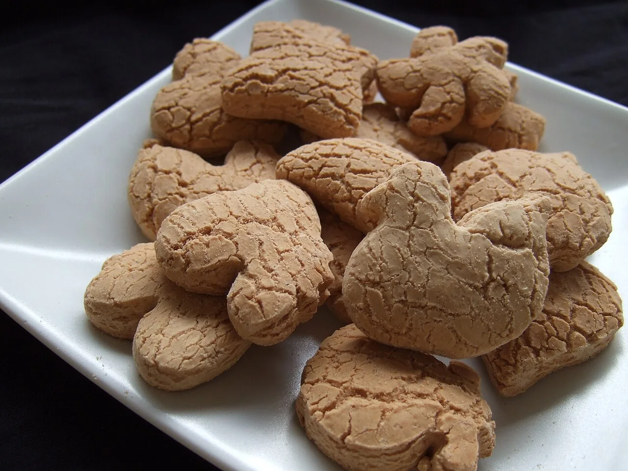Light brown flower-shaped kueh bangkit cookies with cracked surfaces are piled on a white plate.