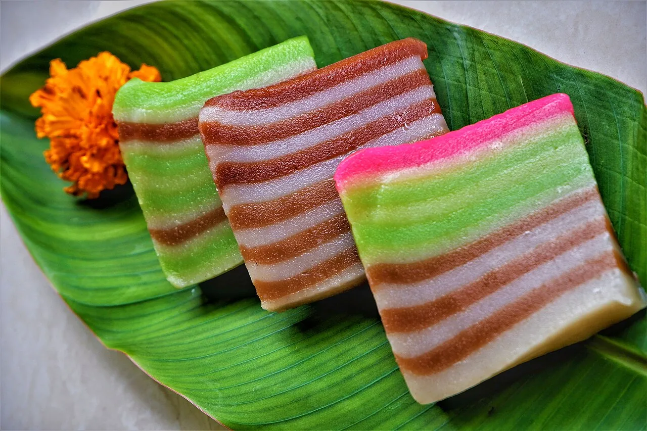 Three slices of striped kueh lapis, colored green, brown, and pink, rest on a green banana leaf beside an orange flower.
